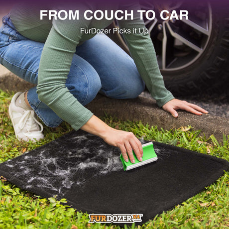 A person kneels on grass next to a car tire, using a FurDozer pet hair remover on a black car mat covered with white fur and debris. Text at the top reads 'FROM COUCH TO CAR FurDozer Picks it Up' and the FurDozer logo appears at the bottom.